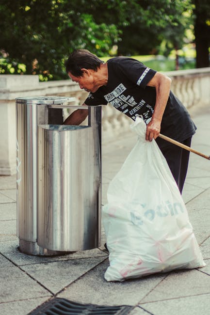 A clear cylindrical waste bin made of lightweight plastic with a mesh-like design, placed on a brown, textured carpeted floor. Inside the bin, there are several crumpled sheets of white paper, some of