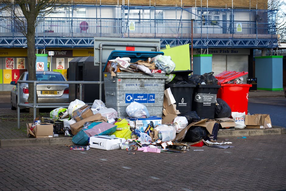 A four-compartment waste disposal bin situated on a train station platform next to railway tracks. The bin is made of stainless steel with a matte finish and is divided into four sections, each design